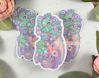 May include: Three holographic stickers featuring a green, purple, and pink cartoon creature with multiple arms and legs. The creature is wearing a purple and pink outfit with a pink and white hat. The stickers are on a white background.