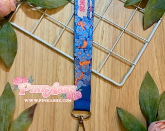 May include: A fabric lanyard featuring a repeating pattern of blue flowers and orange curved shapes on a light blue background. The top section displays the text "R WINGS" in pink letters. A silver metal lobster clasp is attached to the bottom end.