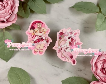 May include: Two pink stickers featuring deer-like characters with floral and heart accents. One deer wears a dress and headphones, the other holds a bow and arrow. The stickers have the text "Pink Ashe" and "www.pinkashe.com".