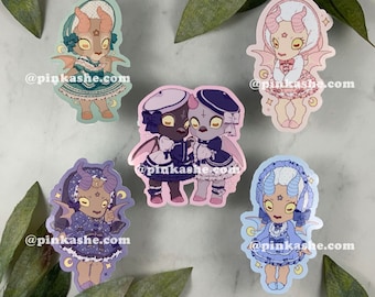 May include: Five colorful stickers featuring cartoon characters with horns and wings. Each sticker has a different color scheme and features a character wearing a dress and holding a small crescent moon. The text '@pinkashe.com' appears on each sticker.