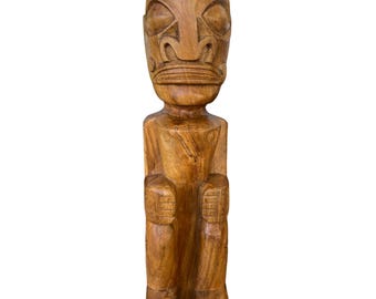 May include: A carved wooden statue of a seated figure with a large head and stylized facial features. The statue is a warm brown color and appears to be made of a single piece of wood. The figure has a rectangular body and is seated with its knees bent.