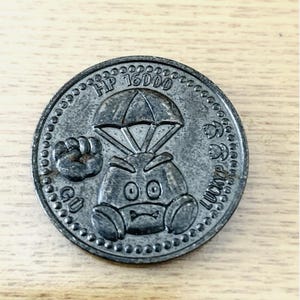 Puede incluir: Una moneda circular de metal gris oscuro con un personaje en relieve con paracaídas, un puño levantado y el texto HP 16000, GU y LUCKYP. La moneda tiene un borde con diseño de cuentas y un acabado texturizado y desgastado en su superficie.