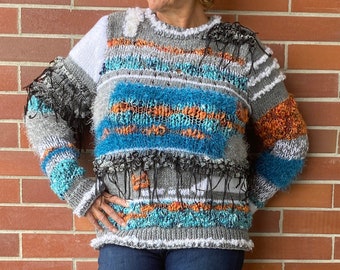 Puede incluir: Un suéter texturizado multicolor con rayas horizontales azules, naranjas y grises. El suéter tiene cuello redondo y se combina con vaqueros azul oscuro y zapatillas blancas. El fondo es una pared de ladrillo rojo.