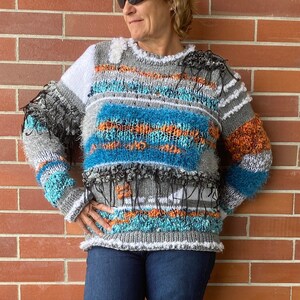 May include: A textured, multi-colored sweater with blue, orange, and gray horizontal stripes. The sweater has a crew neck and is paired with dark blue jeans and white sneakers. The background is a red brick wall.