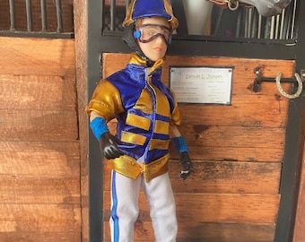Puede incluir: Un traje de jockey azul y dorado con pantalones blancos y un casco azul con visera. El jockey está de pie frente a una puerta de establo de madera con un letrero que dice "Samuel Le Jumper".
