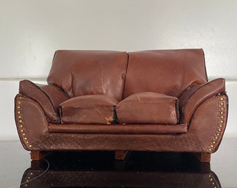 Puede incluir: Un pequeño sofá de cuero marrón con tachuelas doradas alrededor de los bordes. El sofá tiene un aspecto desgastado y está sobre una superficie negra.