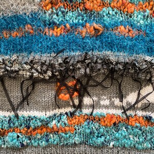 May include: Close-up of a textured textile with a mix of colors and materials. The design features bands of blue, orange, gray, and white, with a variety of textures including fluffy yarn and ribbon accents. The overall effect is a vibrant and tactile piece.