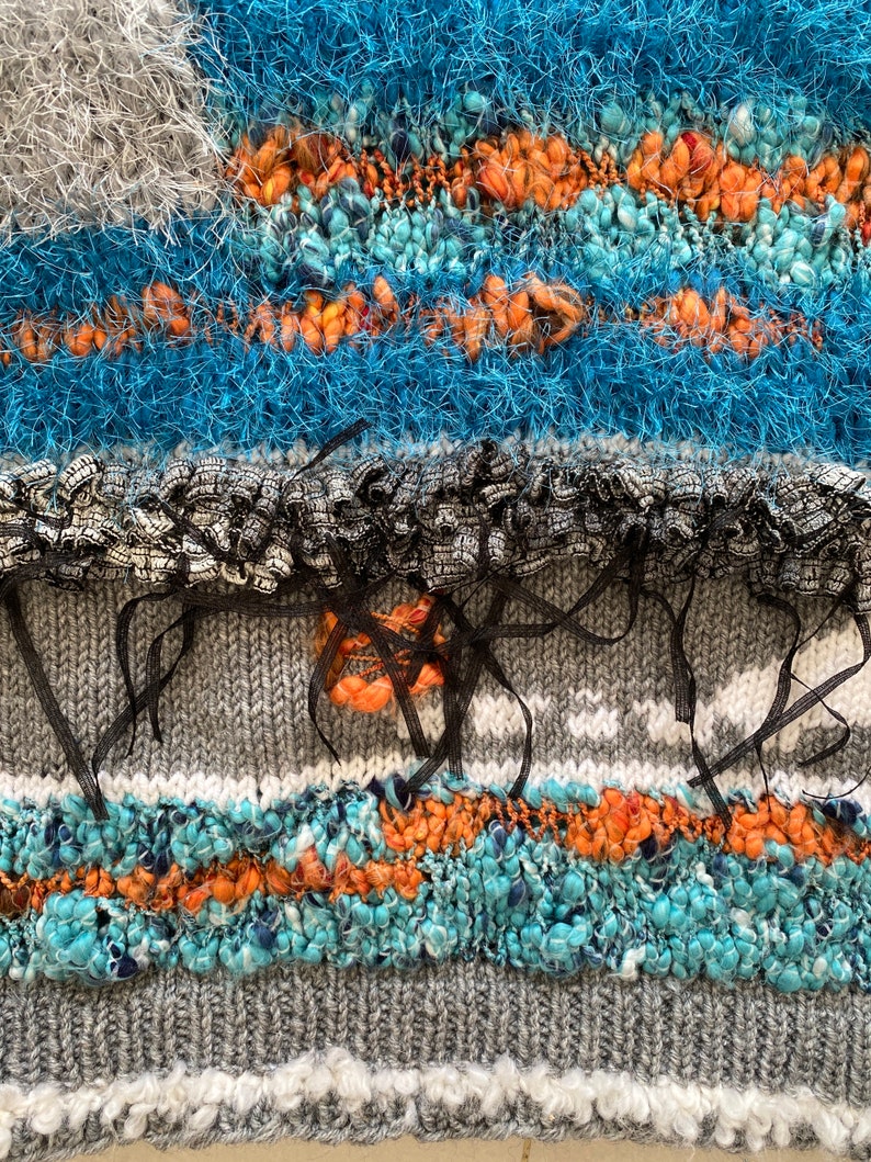May include: Close-up of a textured textile with a mix of colors and materials. The design features bands of blue, orange, gray, and white, with a variety of textures including fluffy yarn and ribbon accents. The overall effect is a vibrant and tactile piece.