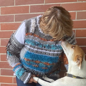 May include: A handmade, multi-colored sweater with a unique, textured design. The sweater features shades of gray, white, blue, and orange, with fringe and textured yarn details. The sweater is a cozy, artistic piece of wearable art.