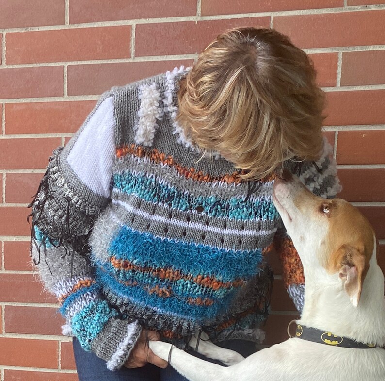 May include: A handmade, multi-colored sweater with a unique, textured design. The sweater features shades of gray, white, blue, and orange, with fringe and textured yarn details. The sweater is a cozy, artistic piece of wearable art.