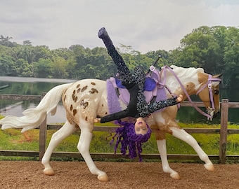 Puede incluir: Un caballo blanco y marrón manchado con una silla de montar y una brida moradas. Una muñeca con cabello morado está boca abajo en el caballo, vestida con un atuendo negro y morado.