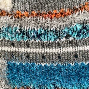 May include: Close-up of a textured knit fabric with a mix of gray, white, orange, and blue yarns. The design includes horizontal stripes and textured embellishments, creating a unique and tactile surface. The knit has a mix of textures.