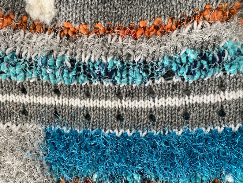May include: Close-up of a textured knit fabric with a mix of gray, white, orange, and blue yarns. The design includes horizontal stripes and textured embellishments, creating a unique and tactile surface. The knit has a mix of textures.