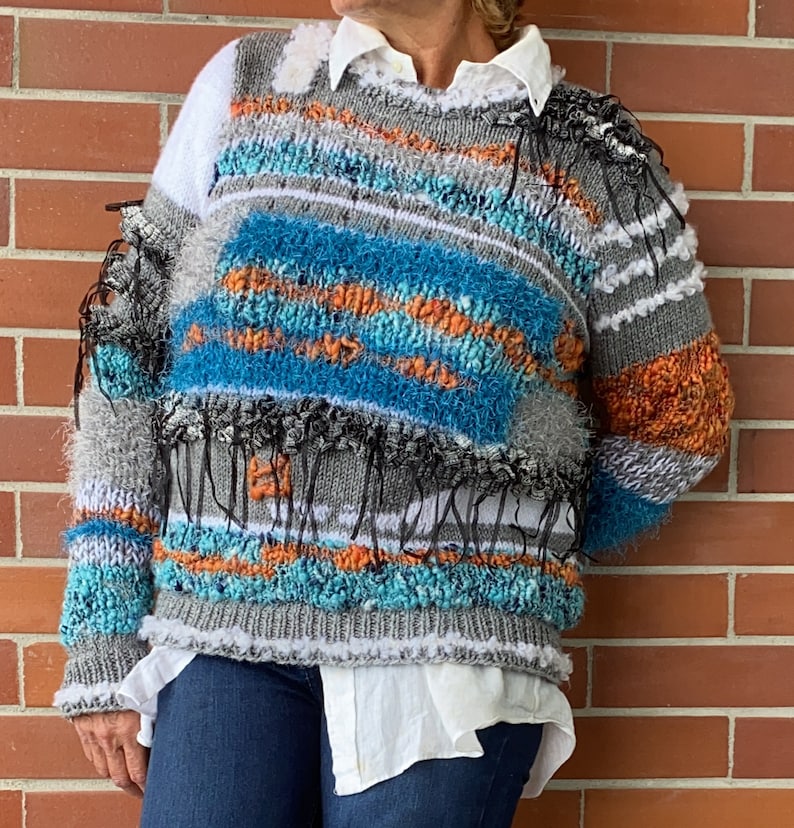 May include: A colorful, textured sweater with a unique design. The sweater features a mix of blue, orange, gray, and white yarns, with fringe and textured details. The sweater is worn over a white collared shirt and blue jeans.