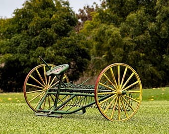 Puede incluir: Un implemento agrícola vintage verde y amarillo con un asiento de madera y ruedas de radios. El implemento está diseñado para rastrillar heno u otros materiales agrícolas.