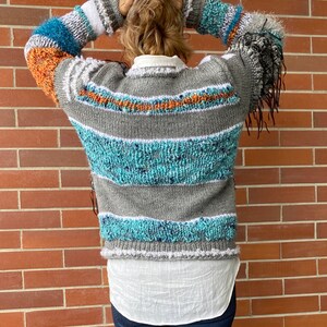 May include: A gray, blue, and orange striped sweater with fringe detail on the sleeves. The sweater has a white collar and is worn over a white shirt. The sweater's design features textured knit sections and horizontal stripes.