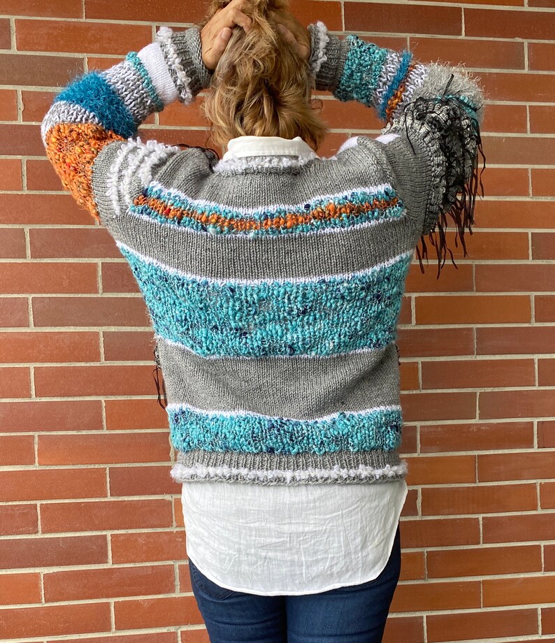 May include: A gray, blue, and orange striped sweater with fringe detail on the sleeves. The sweater has a white collar and is worn over a white shirt. The sweater's design features textured knit sections and horizontal stripes.