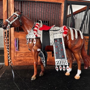 May include: A brown horse model with a silver and red saddle and bridle. The saddle has a black leather seat and the bridle has a red and white braided trim. The horse is wearing a silver breast collar with red jewels. The saddle is decorated with silver plates and the horse's name, "Breyer", is written on the saddle flap.