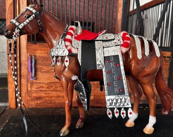 Puede incluir: Un modelo de caballo marrón con una silla de montar y una brida plateadas y rojas. La silla de montar tiene un asiento de cuero negro y la brida tiene un ribete trenzado rojo y blanco. El caballo lleva un collar de pecho plateado con joyas rojas. La silla de montar está decorada con placas plateadas y el nombre del caballo, "Breyer", está escrito en la falda de la silla.