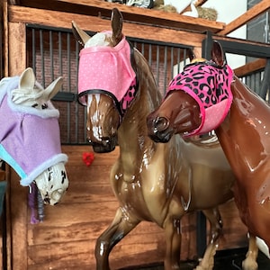 May include: Three toy horses wearing fly masks. The horse on the left has a purple fly mask, the horse in the middle has a pink fly mask with white polka dots, and the horse on the right has a pink fly mask with a black leopard print.