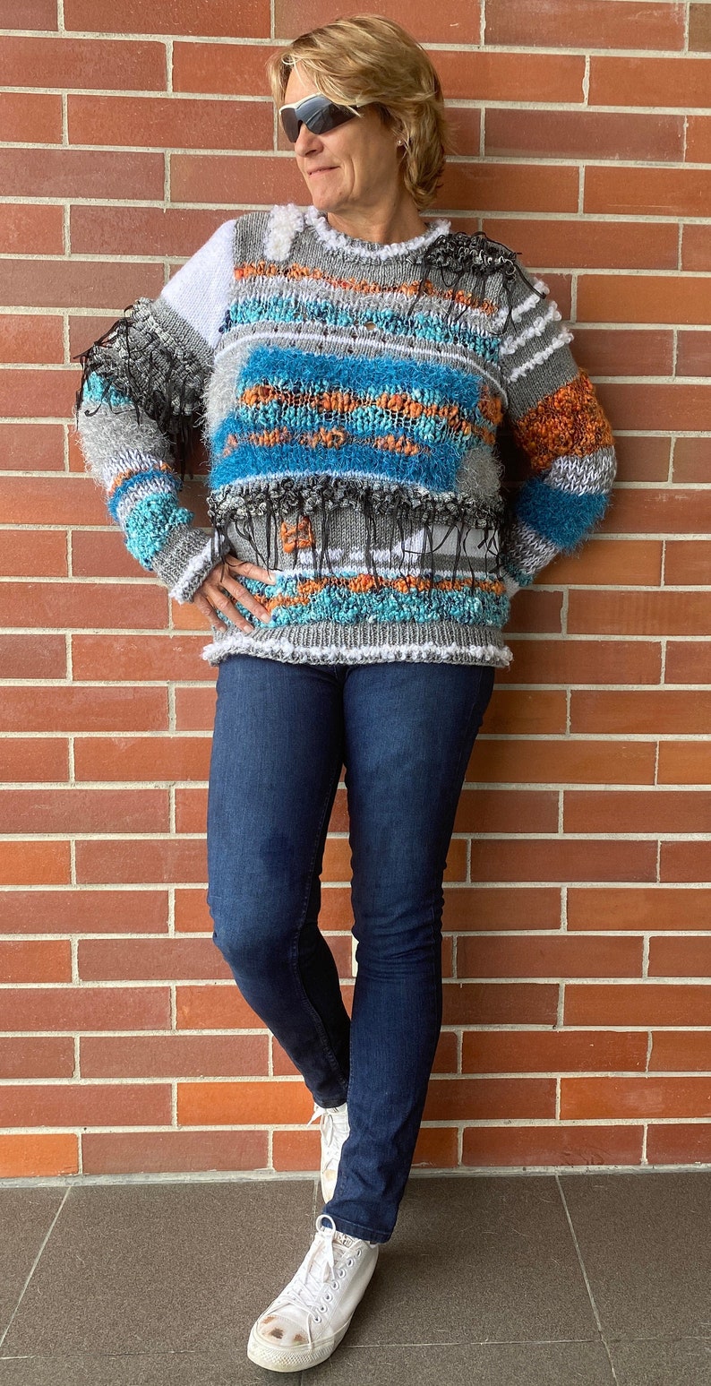 May include: A textured, multi-colored sweater with blue, orange, and gray horizontal stripes. The sweater has a crew neck and is paired with dark blue jeans and white sneakers. The background is a red brick wall.