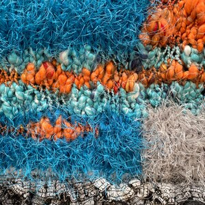 May include: Close-up of a textured textile with a mix of colors and textures. The design features sections of teal, orange, and blue yarn, with a gray knitted background and a black fringe at the bottom. The yarn has a fluffy, textured appearance.
