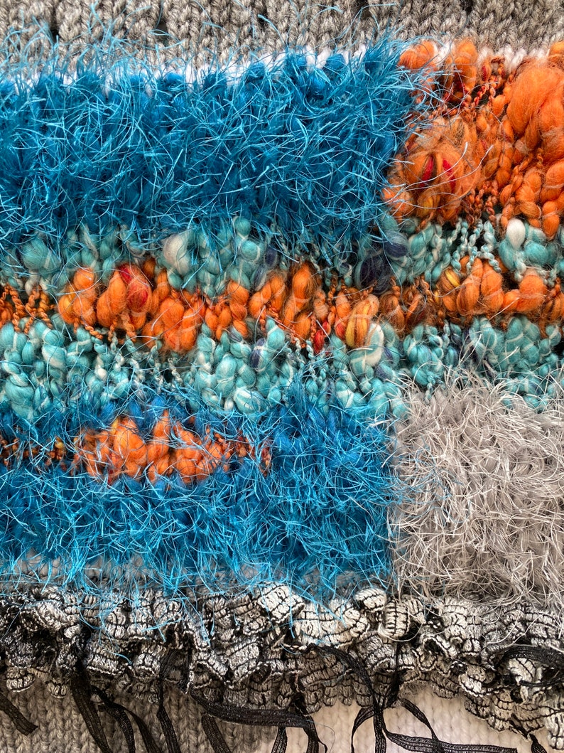 May include: Close-up of a textured textile with a mix of colors and textures. The design features sections of teal, orange, and blue yarn, with a gray knitted background and a black fringe at the bottom. The yarn has a fluffy, textured appearance.