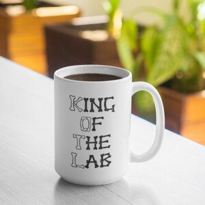 Puede incluir: Taza de café de cerámica blanca con un diseño negro que dice "KING OF THE LAB" con letras en forma de hueso.