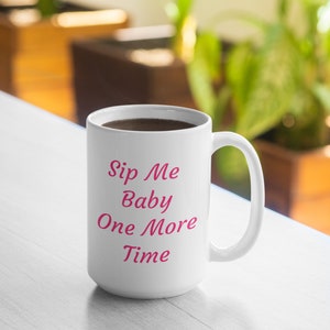 Peut inclure: Une tasse à café en céramique blanche avec une inscription rose qui dit "Sip Me Baby One More Time". La tasse est remplie de café et repose sur une surface en bois clair.