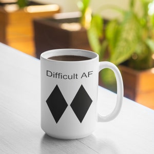 May include: White ceramic coffee mug with black text that reads "Difficult AF" and two black diamond symbols.