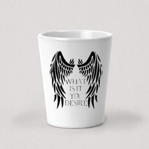 Pode incluir: Um copo de shot de cerâmica branca com uma silhueta preta de asas de anjo e o texto "What is it you desire".