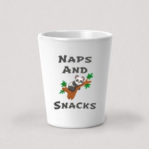 Może przedstawiać: Biały kubek ceramiczny z czarno-białym pandą śpiącym na gałęzi z zielonymi liśćmi. Na kubku znajduje się napis "NAPS AND SNACKS".