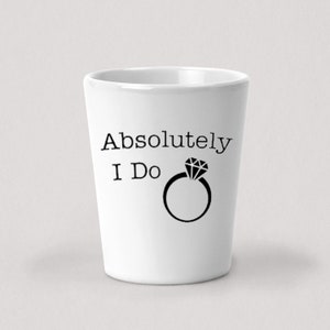 Puede incluir: Un vaso de chupito de cerámica blanca con texto negro que dice "Absolutamente lo hago" y un anillo negro con un diamante.