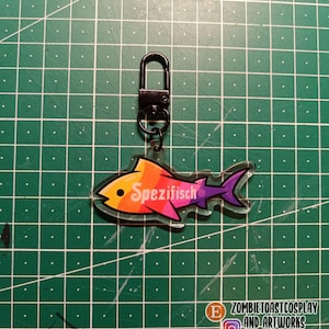 Könnte beinhalten: Ein Schlüsselanhänger aus klarem Acryl mit einem stilisierten Fisch in einem lebendigen Farbverlauf aus Gelb, Orange, Pink und Violett. Das Wort Spezifisch ist in weißer Schreibschrift auf den Fischkörper gedruckt. Mit schwarzem Metallkarabiner.