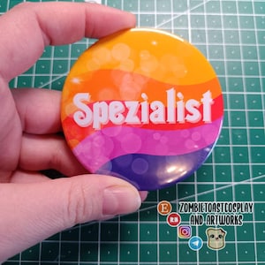 Könnte beinhalten: Ein runder Button mit einem farbenfrohen Farbverlauf, der von Gelb über Orange, Rot, Pink bis Lila reicht. Das Wort "Spezialist" ist in Weiß geschrieben. Der Button wird von einer Hand gehalten.