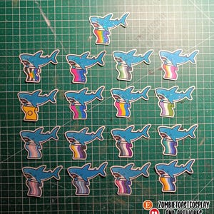Könnte beinhalten: Eine Sammlung blauer Hai-Aufkleber, jeder mit einem Regenbogen-Design. Die Aufkleber zeigen einen Cartoon-Hai mit weißem Bauch und offenem Maul. Die Aufkleber sind auf einer grünen Schneidematte angeordnet.