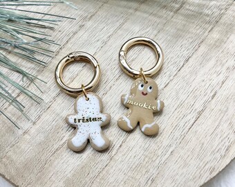 May include: Two gingerbread man charms with gold-coloured key rings. One charm is white with the name "tristan" in black. The other is tan with the word "mookie" in white. The charms are on a wooden surface.