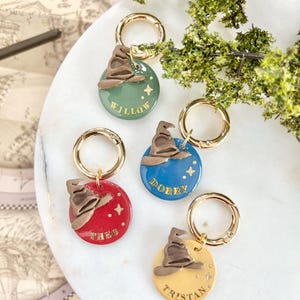 May include: Four gold-toned dog tags with a round, colorful design. Each tag features a brown sorting hat, a name, and gold stars. The names on the tags are Willow, Dobby, Theo, and Tristan.