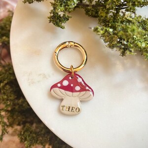 Puede incluir: Encanto de oro con una seta roja y blanca con puntos blancos y el nombre "THEO" escrito en la parte inferior.