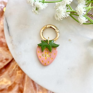 May include: A strawberry-shaped charm with the name "KIKI" in gold lettering. The charm has a pink body, green leaves, and a gold clasp. The background includes white flowers and a marble surface.