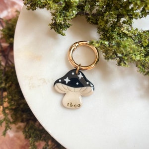 Puede incluir: Un amuleto de cer&aacute;mica en forma de hongo negro y blanco con estrellas doradas y la palabra "theo" escrita en la parte inferior. El amuleto est&aacute; unido a un anillo de metal dorado.