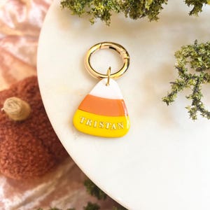 Può includere: Un ciondolo a forma di caramella candy corn con strisce orizzontali bianche, arancioni e gialle. La sezione gialla presenta il nome TRISTAN in lettere maiuscole bianche. Il ciondolo è fissato a un moschettone circolare in metallo dorato.