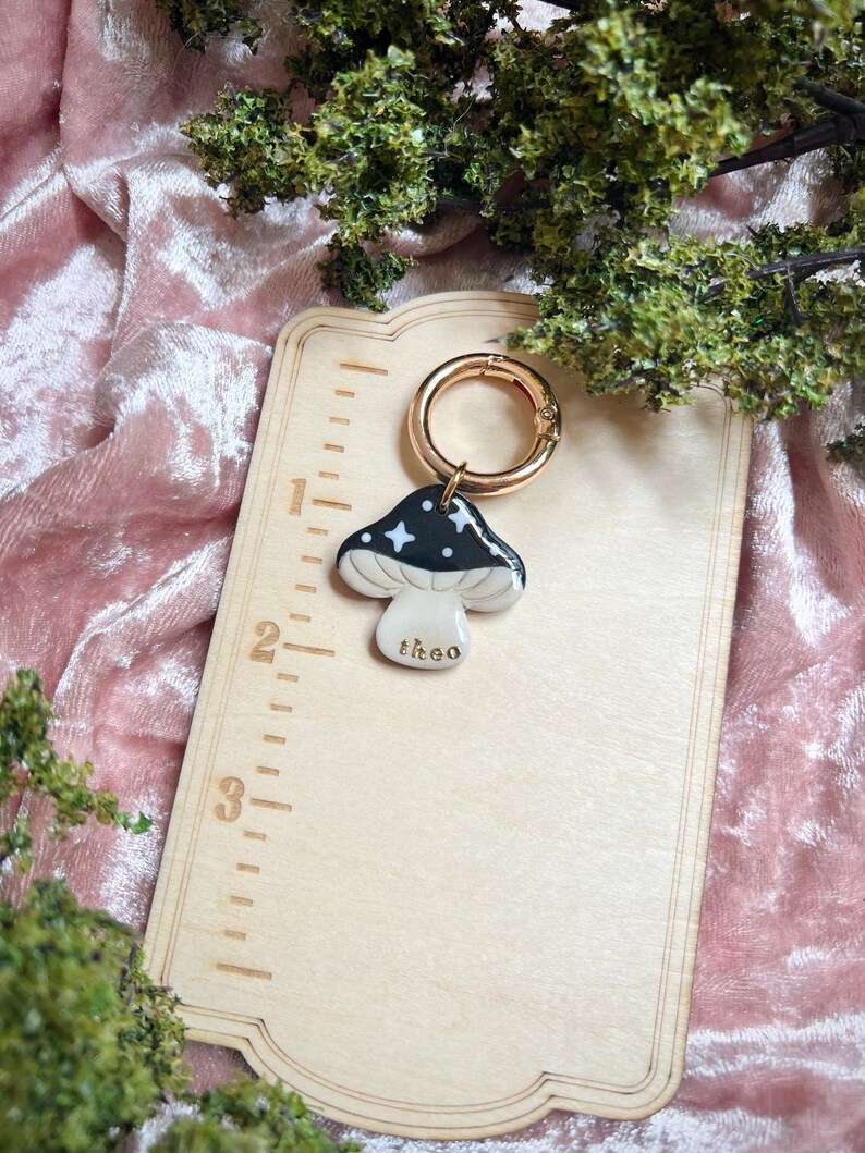 Puede incluir: Un amuleto de hongo negro y blanco con estrellas doradas y la palabra "theo" escrita en &eacute;l. El amuleto est&aacute; unido a un anillo de metal dorado.