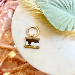 Puede incluir: Un llavero dorado con un dije en forma de s'more. El dije tiene una capa de chocolate marrón, una capa de malvavisco blanco y una capa de galleta Graham. La palabra "THEO" está impresa en la capa inferior.