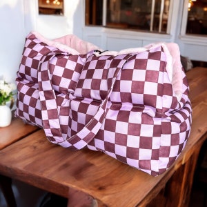 May include: A purple and brown checkered pet bed with a plush pink lining. The bed has two handles and is made of a quilted fabric.