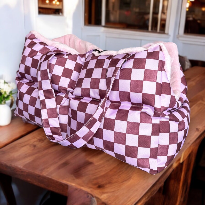 May include: A purple and brown checkered pet bed with a plush pink lining. The bed has two handles and is made of a quilted fabric.