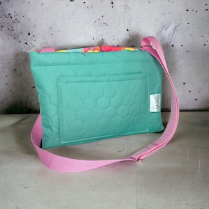 Puede incluir: Un bolso bandolera acolchado de color turquesa con una correa rosa y un pequeño bolsillo en la parte delantera. El bolso tiene una etiqueta blanca con la palabra "Gina" escrita en ella.