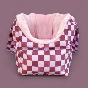 May include: A pink and brown checkered pet bed with a soft pink plush lining. The bed has a rounded shape and two handles on the sides.
