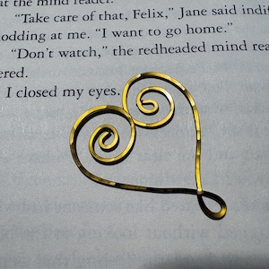 May include: A gold-colored wire heart ornament featuring two symmetrical spiral coils at the top and a pointed base. The heart is placed on a page of a book with printed text that reads: Take care of that, Felix, I want to go home, Don't watch, I closed my eyes.