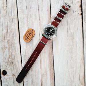 May include: A brown leather watch strap with white stitching and a silver watch with a black face. The strap has the text "Swag Bridge Leather Straps" on a small leather tag.